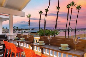 Eine Terrasse mit Holztischen und orangefarbenen Stühlen mit Blick auf einen Yachthafen, wo Boote und Palmen zu sehen sind.