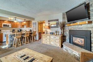 Ein Wohnzimmer mit Kamin, einem an der Wand befestigten Fernseher, Holzmöbeln und einer Küchenzeile mit Stein Arbeitsplatten.