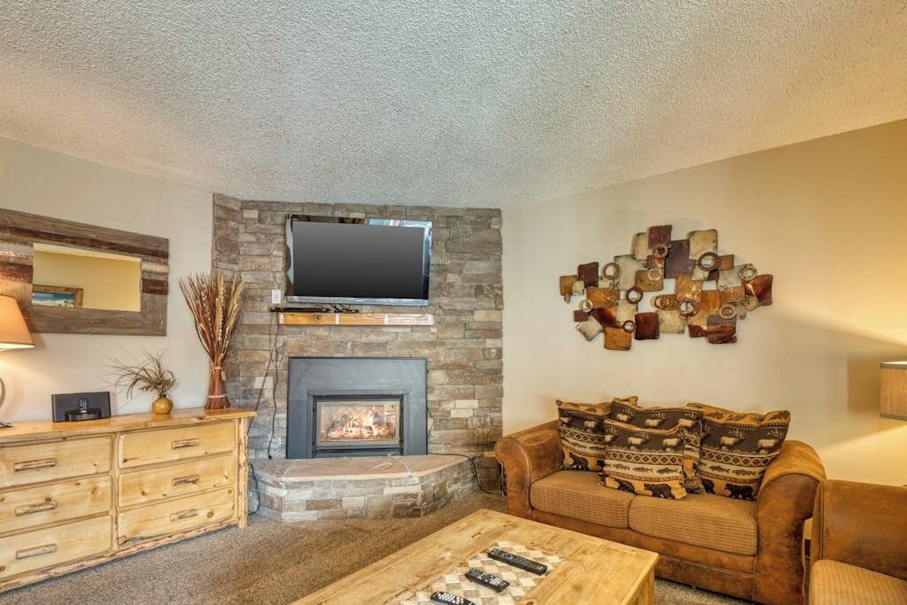 A living room with a stone fireplace, a flat-screen TV mounted above it, a wooden coffee table, a brown leather sofa with patterned cushions, and a decorative wall art piece.