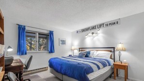 Een slaapkamer met een bed, twee nachtkastjes met lampen, een bureau met stoel, een raam met blauwe gordijnen en een bord op de muur.