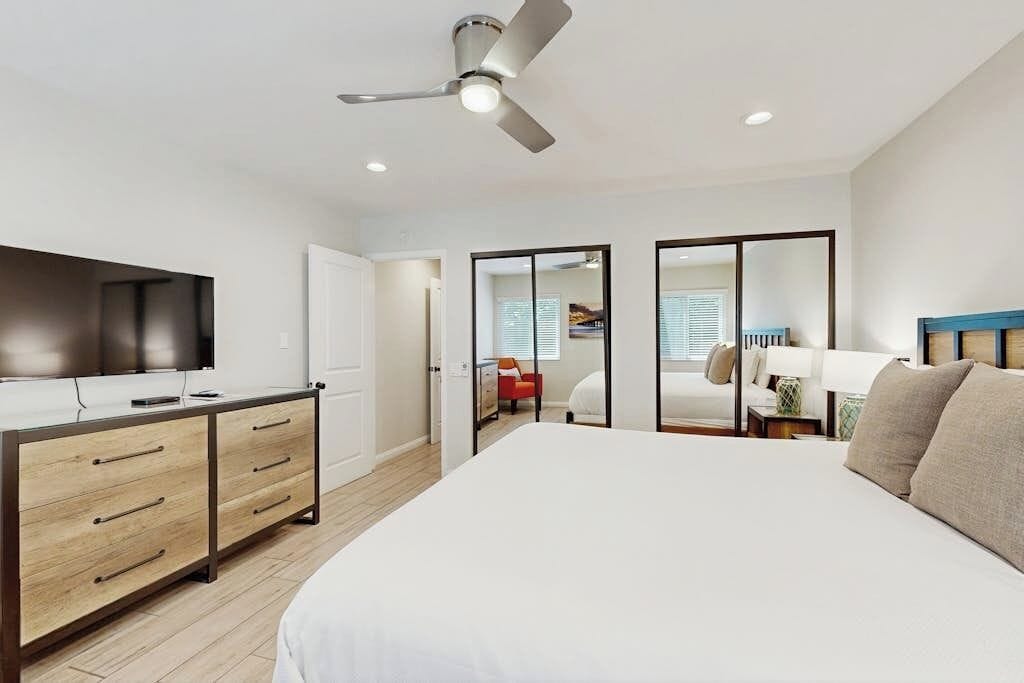 A bedroom with a bed, a desk with drawers, a ceiling fan, and a television.