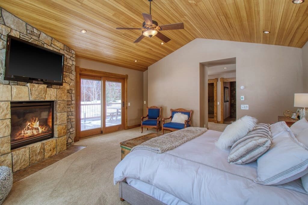 A bedroom with a stone fireplace, a large bed, a ceiling fan, and a TV mounted on the wall.