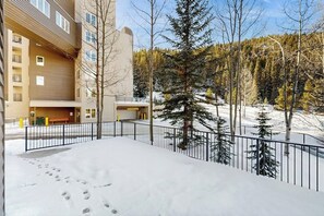 A snow-covered outdoor area with a building, a fenced walkway, and a forested hillside. - Cozy ski-in/out condo with shared hot tub and pool, mountain views (Winter Park)