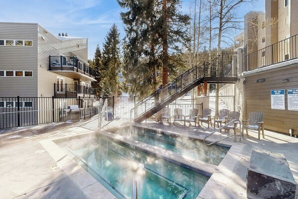 A pool area with a clear view of the pool, a staircase, and surrounding buildings. - Cozy ski-in/out condo with shared hot tub and pool, mountain views (Winter Park)