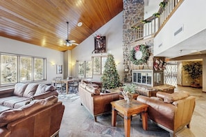 A spacious living area with leather sofas, a stone fireplace, and a large window overlooking a forest. - Cozy ski-in/out condo with shared hot tub and pool, mountain views (Winter Park)