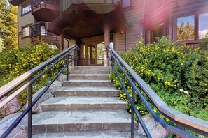 Ein Holzhaus mit einer Treppe, die zu einer Veranda führt, umgeben von Grünpflanzen und gelben Blumen.