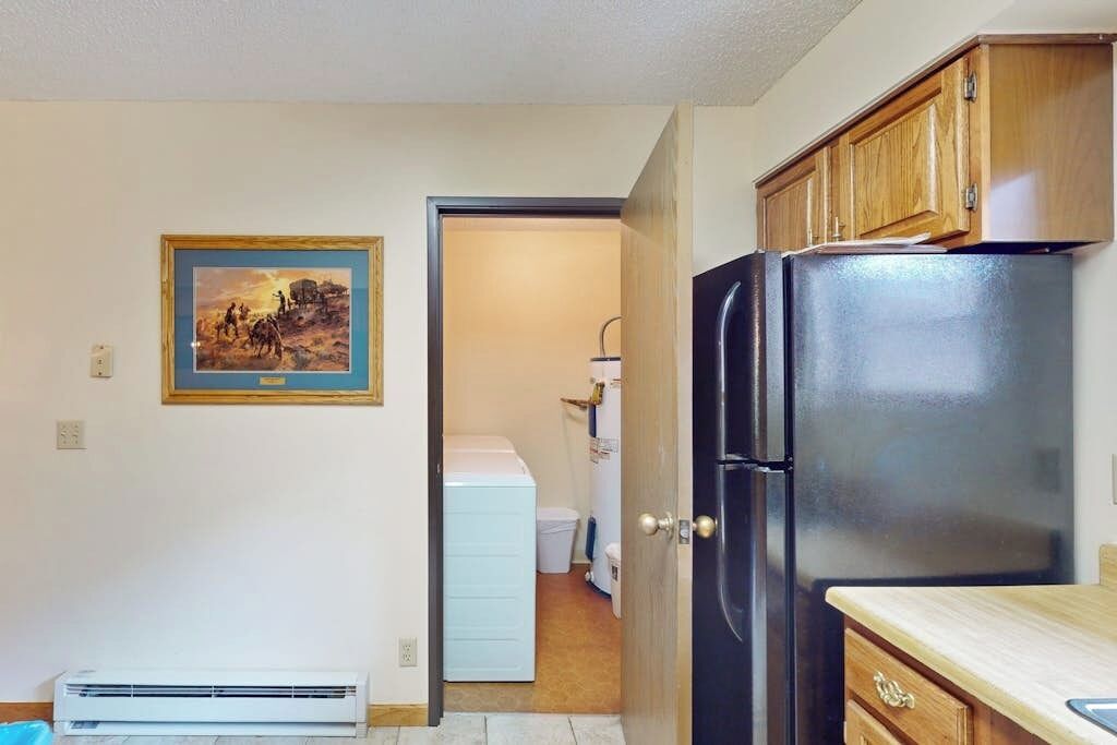 A kitchen with a black refrigerator, wooden cabinets, a white washing machine, and a framed picture on the wall.