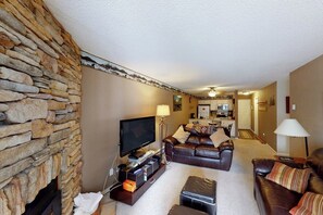 Ein Wohnzimmer mit Steinwand, Fernseher, Ledersofas und einer Küche im Hintergrund.