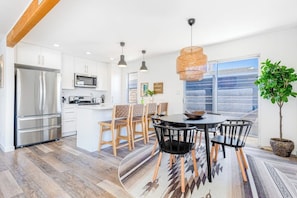A modern kitchen with white cabinets, a stainless steel refrigerator, and a dining area with a round table and chairs. - Charming gem near the beach with large backyard, cornhole, fast WiFi & grill (San Clemente)