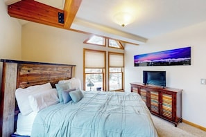 Ein Schlafzimmer mit Bett, Fernseher, Holz-Kopfteil, Fenster und einem Bild an der Wand.
