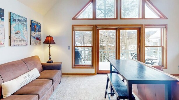 A room with a brown leather sofa, a wooden dining table, and a large window with a view of trees. - Dog-friendly condo right in the center of town - walk to & from skiing (Breckenridge)