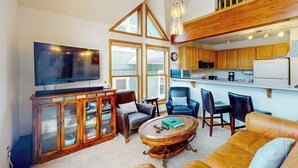 A spacious living room with a television, seating, a coffee table, and a kitchen area with wooden cabinets and a dining table. - Dog-friendly condo right in the center of town - walk to & from skiing (Breckenridge)