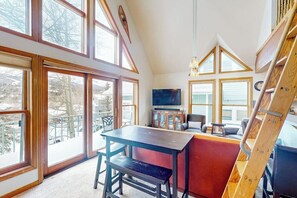 Ein Zimmer mit hohen Decken, großen Fenstern, einem Holztisch, einer Bank und einem Fernseher.