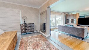 Un vaste salon agrémenté d’un grand tapis, de meubles en bois, d’un téléviseur et d’un coin cuisine visible à travers une porte ouverte.