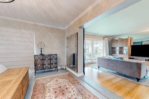 A spacious living room with a large rug, wooden furniture, a TV, and a kitchen area visible through an open door. - Glamorous, dog-friendly condo with pool, hot tub & W/D - snowbird-friendly (Newport Beach)