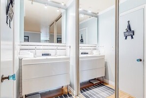 A modern bathroom with two sinks, a large mirror, and a door with a decorative sign. - Glamorous, dog-friendly condo with pool, hot tub & W/D - snowbird-friendly (Newport Beach)