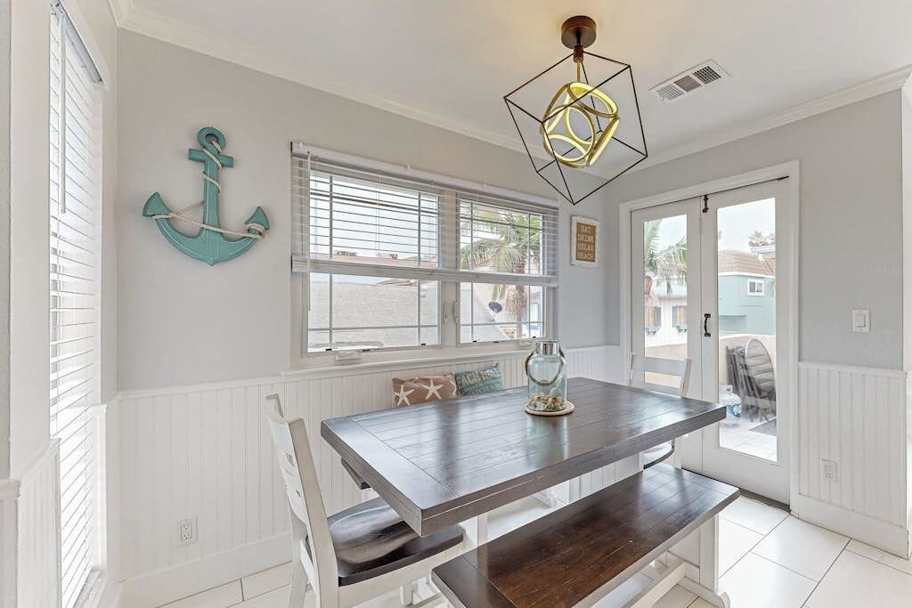 A dining area with a wooden table, white chairs, a window with blinds, and a decorative anchor on the wall.
