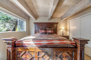 Ein Schlafzimmer mit Holz-Kopfteil, einem Bett mit gemusterten Betttextilien, einem Nachttisch mit Lampe und einem Fenster mit Blick auf Bäume.