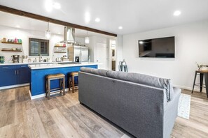 Eine moderne Küche mit blauer Arbeitsplatte, Holzstühlen und einem grauen Sofa in einem Wohnbereich.