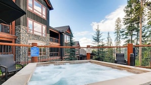 Un jacuzzi en un deck de piedra con barandilla de madera, rodeado por un edificio de exterior de piedra y madera, y un fondo boscoso.
