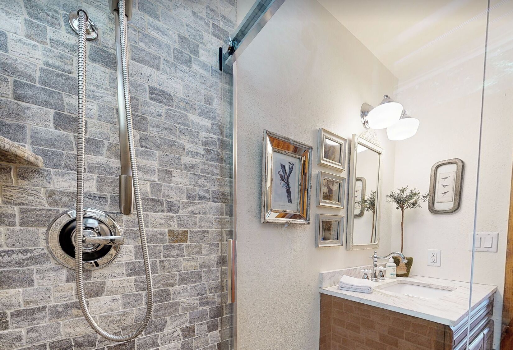 Un baño con pared de piedra, ducha, lavamanos con espejo y cuadros enmarcados en la pared.