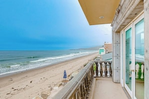 Ein Balkon mit Blick auf Strand und Meer, eine Glastür sowie ein kleiner Tisch mit einem Stuhl.