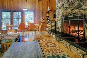 Ein gemütliches Zimmer mit Kamin, Holzwänden, einem Essbereich mit orangefarbenen Stühlen und einem großen Fenster.