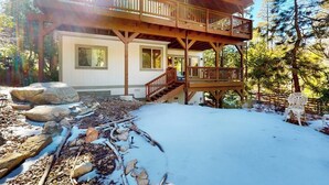 Una cabaña con terraza de madera, porche cubierto y suelo cubierto de nieve.