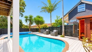 A swimming pool with a wooden deck, outdoor furniture, and a house in the background.