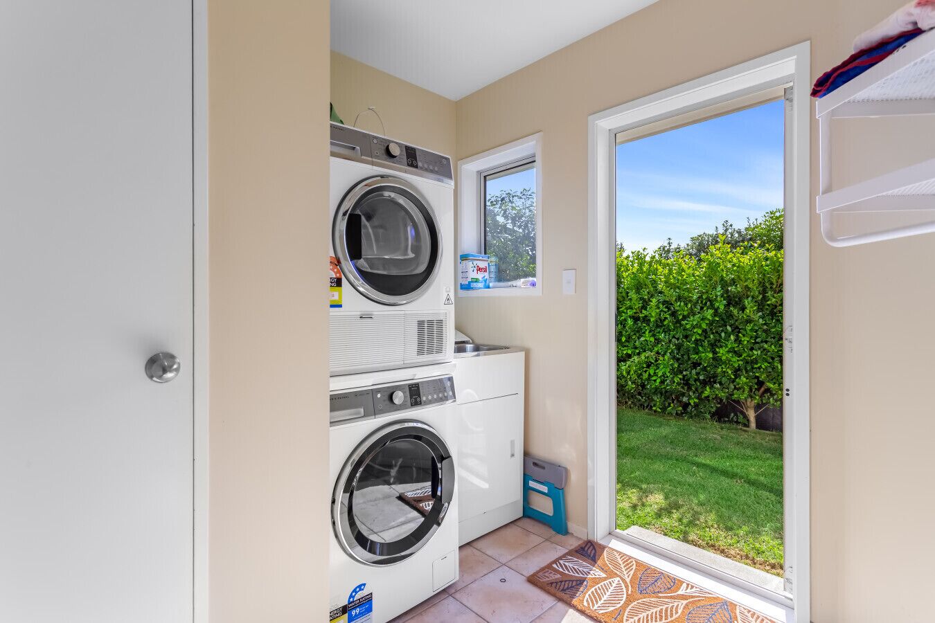 A laundry area with two washing machines, a drying rack, and a window with a view of a green hedge.