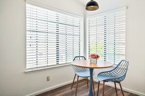 A dining area with a round table and two chairs, large windows with blinds, and a pendant light. - 2-story centrally located home with grill, patio, fireplace, streaming, & W/D (Grover Beach)