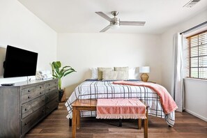 A bedroom with a bed, a ceiling fan, a TV, a dresser, a plant, and a window with blinds.