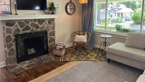 Una sala de estar con chimenea de piedra, un televisor montado encima, un sillón de mimbre, una mesita redonda y vista a una casa a través de una ventana.