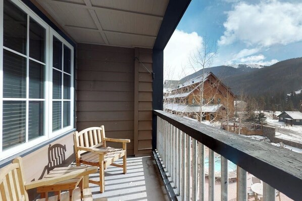 A wooden deck with chairs, a window with blinds, and a view of a mountain lodge. - Cozy Mountain Condo with Pool & Hot Tub - Close to the Slopes (Keystone)