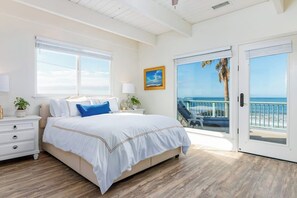 Een slaapkamer met een groot bed, een nachtkastje, een lamp, een ingelijst schilderij en uitzicht op de oceaan door een schuifdeur.