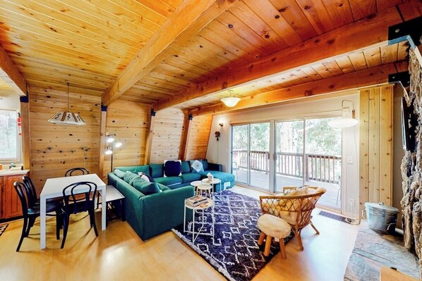 A cozy cabin interior with a wooden ceiling, a green sofa, a dining table with chairs, and a large window. - Dog-friendly 2-story home with large deck, tree views, fireplace, & W/D (Crestline)