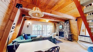 A cozy cabin interior with a stone fireplace, wooden ceiling beams, a dining area, and a large window. - Dog-friendly 2-story home with large deck, tree views, fireplace, & W/D (Crestline)
