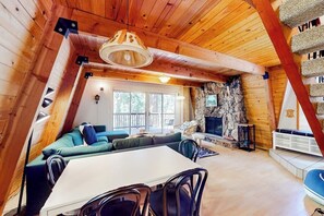 A cozy cabin interior with a stone fireplace, wooden ceiling beams, a dining area, and a large window. - Dog-friendly 2-story home with large deck, tree views, fireplace, & W/D (Crestline)