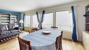 A room with a dining table, chairs, a sofa, a bookshelf, and large windows with curtains. - Classic waterfront beach house with a furnished deck & ocean views (Cayucos)