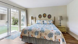 A bedroom with a bed, two bedside tables, a sliding glass door leading to a balcony, and three wall-mounted decorative plates. - Classic waterfront beach house with a furnished deck & ocean views (Cayucos)