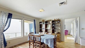 A dining area with a table set for four, wooden chairs, a patterned tablecloth, and a bookshelf with various items. - Classic waterfront beach house with a furnished deck & ocean views (Cayucos)