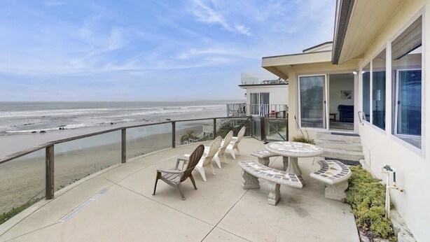 A balcony with a view of the beach, outdoor furniture, and a building with large windows. - Classic waterfront beach house with a furnished deck & ocean views (Cayucos)