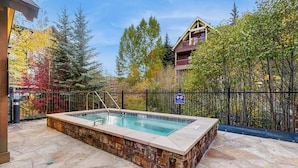 A rectangular hot tub with a stone surround, a metal fence, and trees with autumn foliage in the background. - Spacious, walk-to-lift condo with vaulted ceilings, pool, hot tubs & gym (Breckenridge)