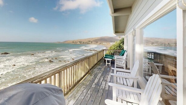 Una terraza de madera con sillas blancas que da a una playa con olas.