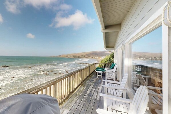 Eine Holzterrasse mit weißen Stühlen mit Blick auf einen Strand und Wellen.