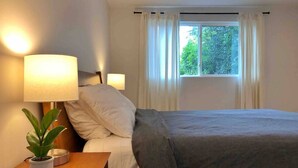 Un dormitori amb llit, taulells laterals, una planta sobre la tauleta i una finestra amb cortines.