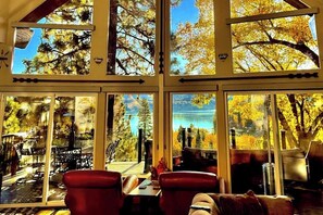 Ein Zimmer mit großen Fenstern, Blick auf Bäume und einen See, sowie einem Essbereich mit Stühlen und Tischen.