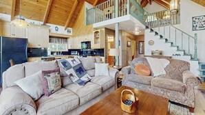 Amplia sala de estar con techo alto, vigas de madera, una escalera y una cocina al fondo.