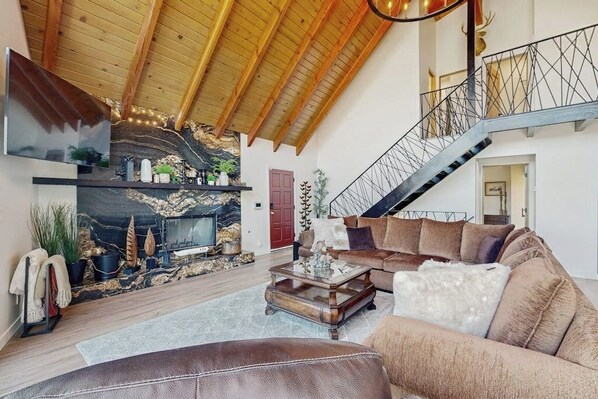 A spacious living area with a fireplace, a sofa, a coffee table, and a staircase. - Modern chalet w/expansive deck, gas grill, & game room - close to lake & town (Lake Arrowhead)