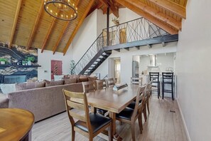 A spacious loft with a high ceiling, a wooden dining table, a staircase, and a cozy living area. - Modern chalet w/expansive deck, gas grill, & game room - close to lake & town (Lake Arrowhead)